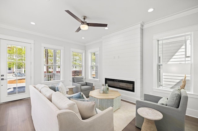 living area with dark wood-type flooring, a ceiling fan, crown molding, and a glass covered fireplace