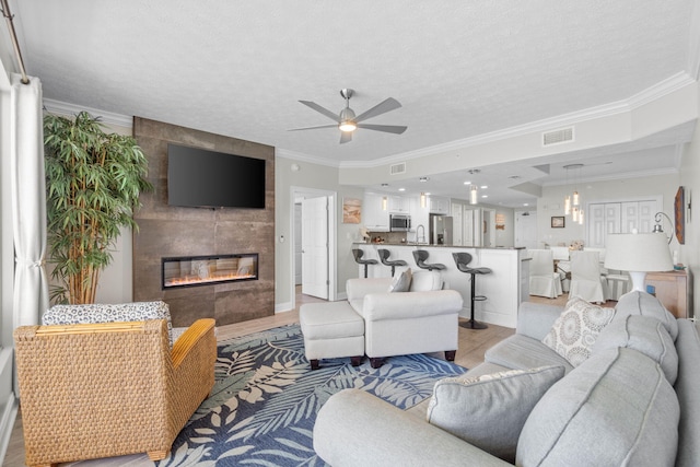 living room featuring a tiled fireplace, visible vents, a ceiling fan, and ornamental molding