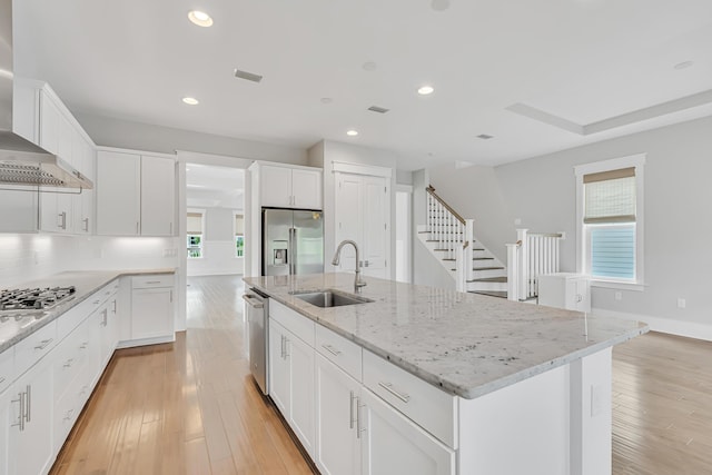 kitchen featuring stainless steel appliances, a sink, a kitchen island with sink, wall chimney exhaust hood, and white cabinetry