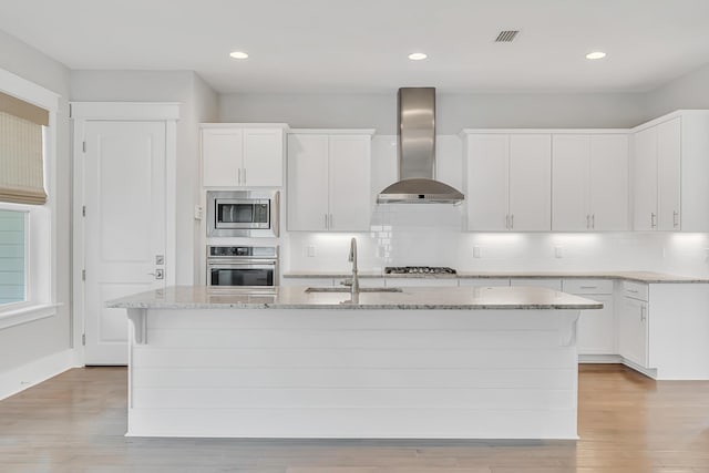 kitchen with stainless steel appliances, a sink, wall chimney range hood, light wood-style flooring, and tasteful backsplash