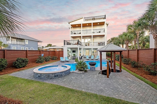 back of house at dusk featuring a balcony, a gazebo, a fenced backyard, a patio, and a pool with connected hot tub