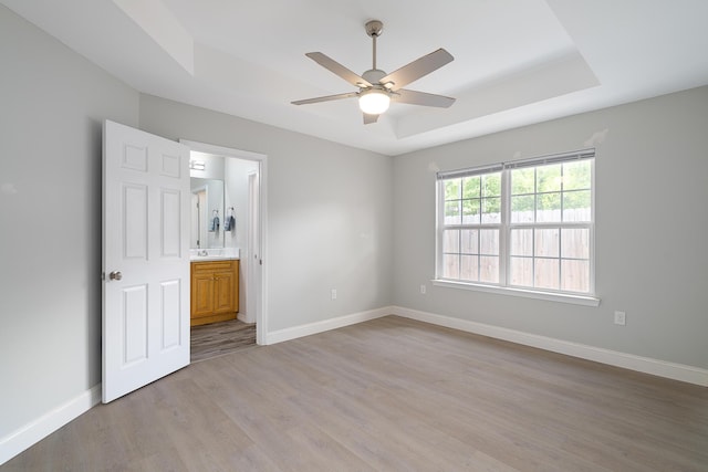 unfurnished bedroom featuring baseboards, ceiling fan, a raised ceiling, light wood finished floors, and ensuite bath