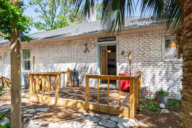 view of exterior entry with a deck, roof with shingles, a chimney, and brick siding