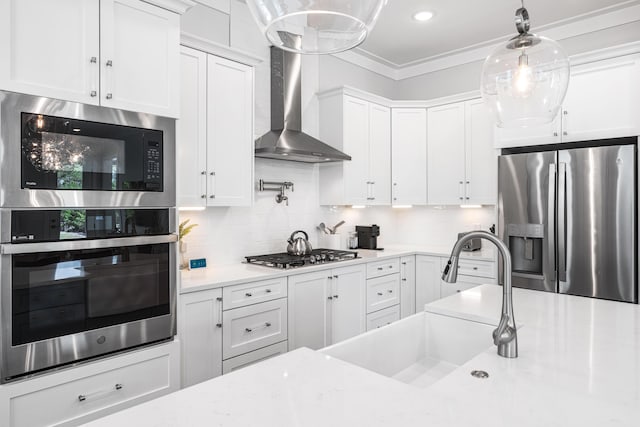 kitchen with stainless steel appliances, a sink, wall chimney range hood, white cabinetry, and ornamental molding