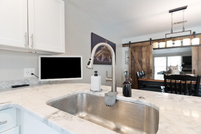 kitchen with light stone countertops, a barn door, decorative light fixtures, a sink, and white cabinetry