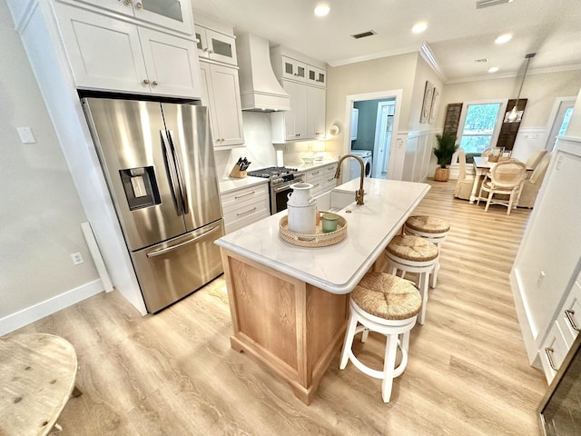 kitchen with custom range hood, stainless steel appliances, crown molding, an island with sink, and light wood-type flooring