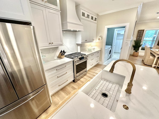 kitchen with custom exhaust hood, stainless steel appliances, ornamental molding, washing machine and clothes dryer, and white cabinets