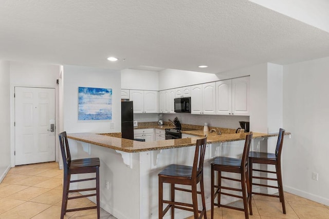kitchen featuring black appliances, a peninsula, light tile patterned flooring, dark stone countertops, and white cabinetry