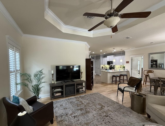 living area with crown molding, visible vents, light wood-style floors, ceiling fan, and a raised ceiling