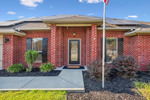 entrance to property with brick siding, roof with shingles, and solar panels