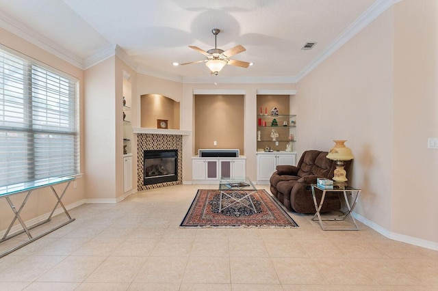 tiled living area featuring built in shelves, a ceiling fan, ornamental molding, a tile fireplace, and baseboards