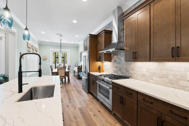 kitchen featuring light wood-style floors, high quality appliances, wall chimney exhaust hood, a sink, and decorative light fixtures