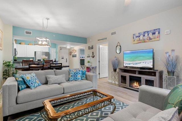 living area with a notable chandelier, a warm lit fireplace, light wood-style flooring, and visible vents