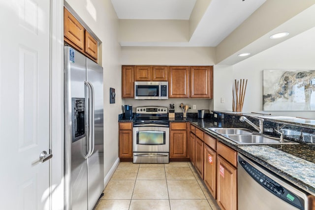 kitchen featuring brown cabinetry, light tile patterned floors, a sink, dark stone counters, and appliances with stainless steel finishes