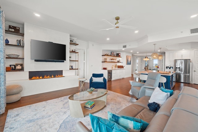 living room with a warm lit fireplace, dark wood-type flooring, recessed lighting, crown molding, and ceiling fan
