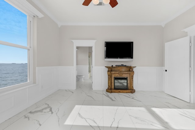 unfurnished living room with a ceiling fan, marble finish floors, wainscoting, a glass covered fireplace, and crown molding