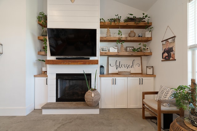 living room featuring a fireplace with raised hearth, vaulted ceiling, concrete flooring, and baseboards