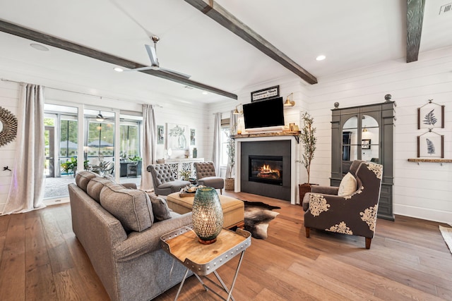 living room featuring ceiling fan, beamed ceiling, wood-type flooring, a glass covered fireplace, and wooden walls