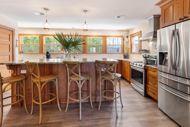kitchen with appliances with stainless steel finishes, wall chimney exhaust hood, brown cabinetry, and wood finished floors
