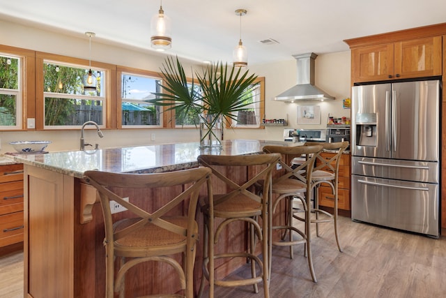 kitchen with stainless steel fridge with ice dispenser, wall chimney exhaust hood, light wood-type flooring, and light stone countertops