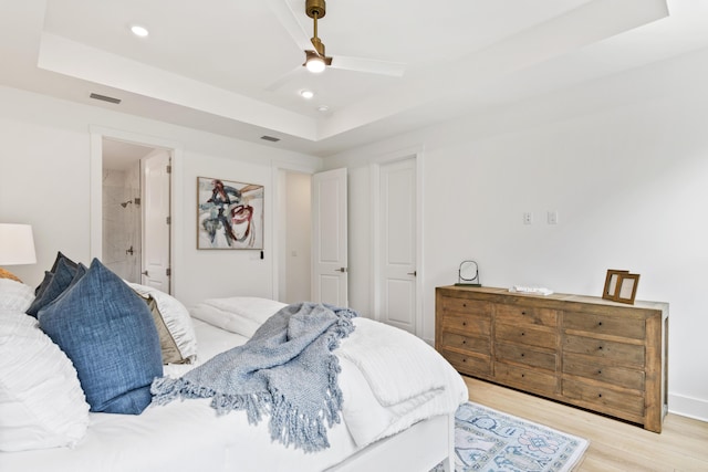 bedroom with a tray ceiling, light wood-type flooring, recessed lighting, a ceiling fan, and connected bathroom