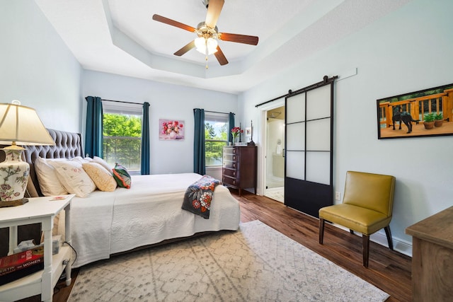 bedroom featuring a barn door, a raised ceiling, wood finished floors, a ceiling fan, and ensuite bath