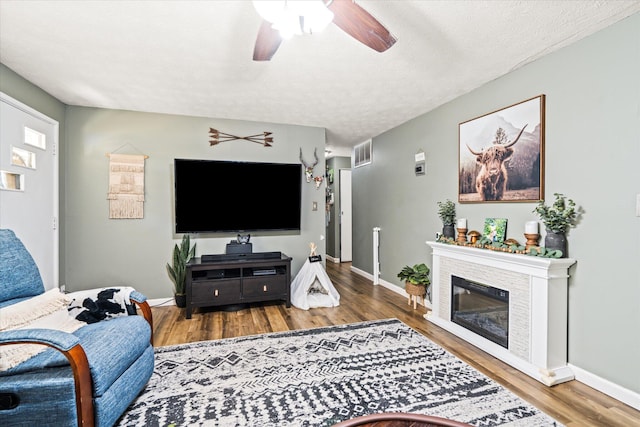 living room featuring visible vents, a ceiling fan, a glass covered fireplace, wood finished floors, and baseboards