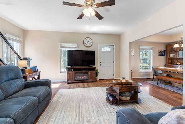 living room featuring a glass covered fireplace, ceiling fan with notable chandelier, wood finished floors, and baseboards