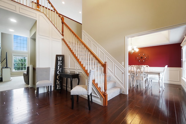 staircase with a decorative wall, a high ceiling, an inviting chandelier, wainscoting, and wood finished floors