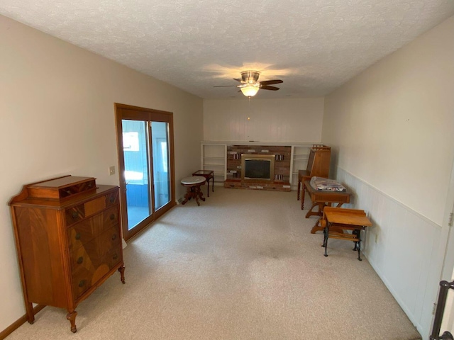 living room featuring a ceiling fan, light carpet, wainscoting, a textured ceiling, and a brick fireplace