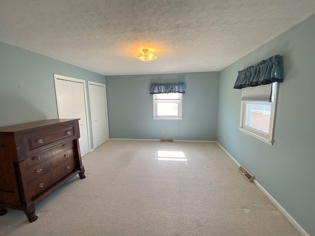 unfurnished bedroom featuring baseboards, visible vents, a textured ceiling, light colored carpet, and two closets