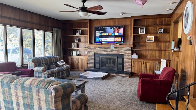 carpeted living area featuring built in shelves, wooden walls, a ceiling fan, ornamental molding, and a brick fireplace