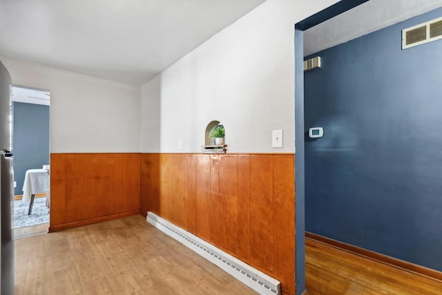 interior space with a wainscoted wall, light wood finished floors, a baseboard radiator, visible vents, and wooden walls