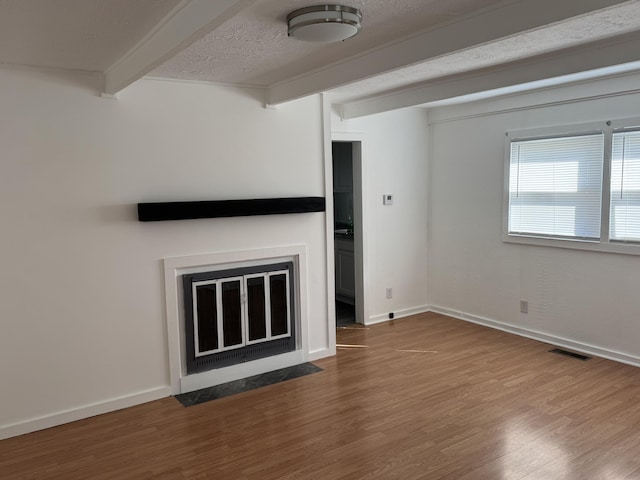 unfurnished living room with a textured ceiling, wood finished floors, a fireplace with flush hearth, baseboards, and beam ceiling