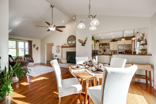 dining space with a fireplace, light wood-style floors, high vaulted ceiling, recessed lighting, and ceiling fan with notable chandelier