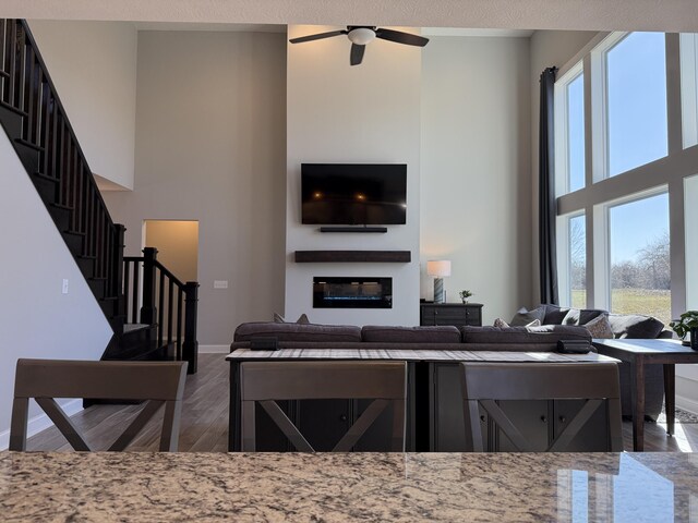 living area featuring stairs, a towering ceiling, wood finished floors, ceiling fan, and a glass covered fireplace