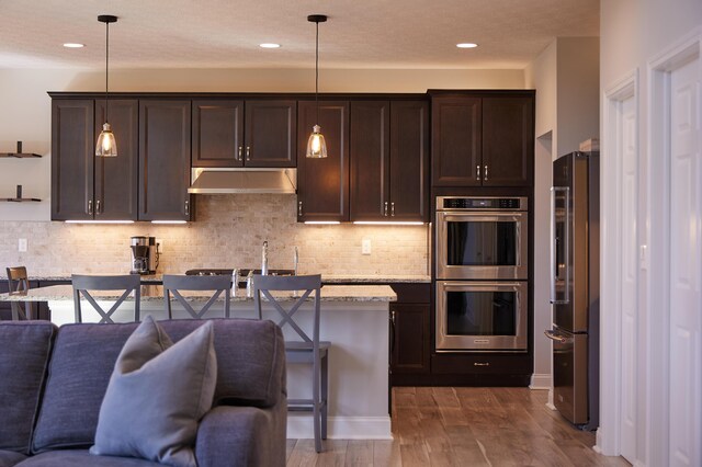 kitchen with decorative backsplash, stainless steel appliances, dark brown cabinetry, under cabinet range hood, and open floor plan