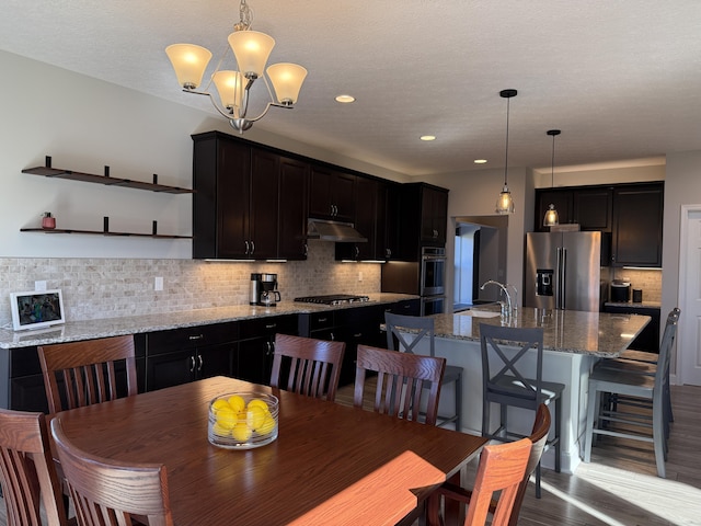 kitchen featuring decorative backsplash, a kitchen island with sink, stainless steel appliances, under cabinet range hood, and a sink
