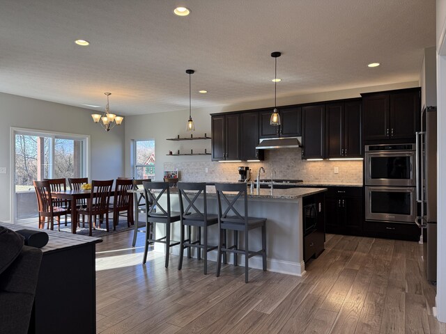 kitchen featuring black microwave, stainless steel double oven, dark wood finished floors, under cabinet range hood, and decorative backsplash