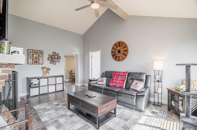 living room featuring a ceiling fan, high vaulted ceiling, beam ceiling, a fireplace with raised hearth, and arched walkways