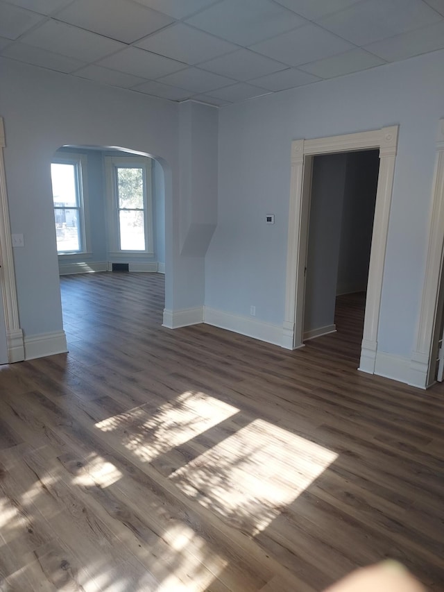 empty room with a drop ceiling, arched walkways, baseboards, and dark wood-style floors