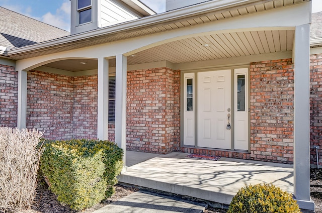 entrance to property featuring covered porch, a shingled roof, and brick siding