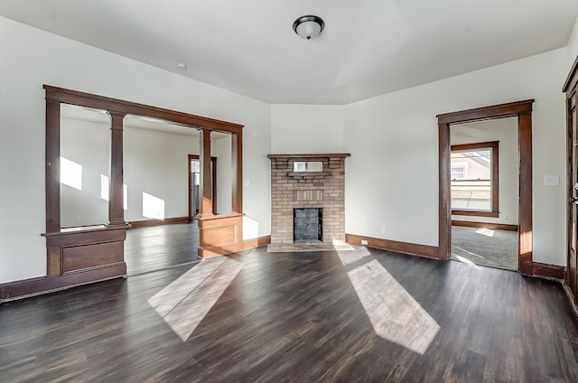 unfurnished living room with dark wood-style floors, a brick fireplace, baseboards, and ornate columns