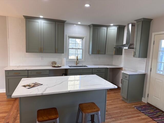 kitchen with wall chimney range hood, gray cabinetry, a sink, a kitchen breakfast bar, and backsplash