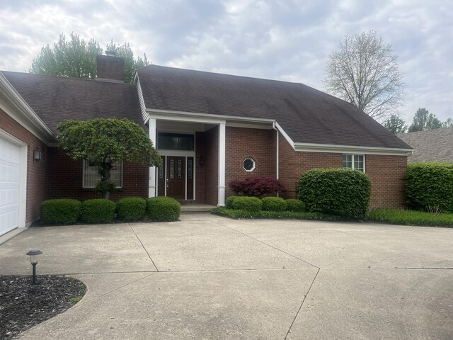 single story home featuring a shingled roof, brick siding, a garage, a chimney, and concrete driveway