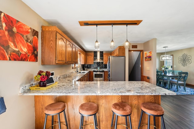 kitchen featuring wall chimney exhaust hood, stainless steel appliances, a peninsula, brown cabinets, and dark wood-type flooring