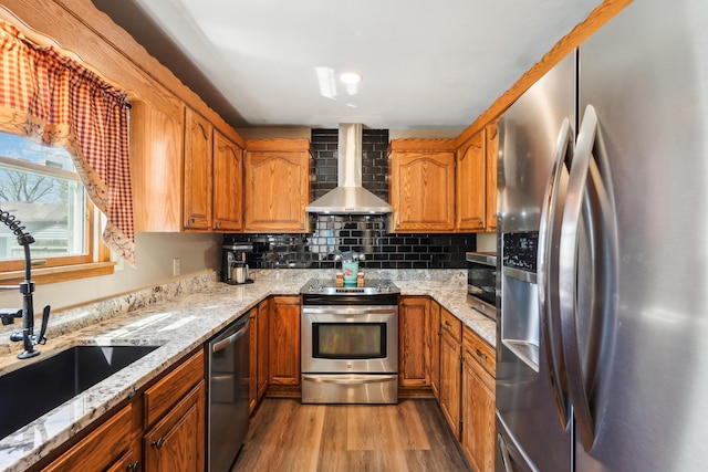 kitchen featuring wall chimney exhaust hood, stainless steel appliances, a sink, wood finished floors, and brown cabinetry