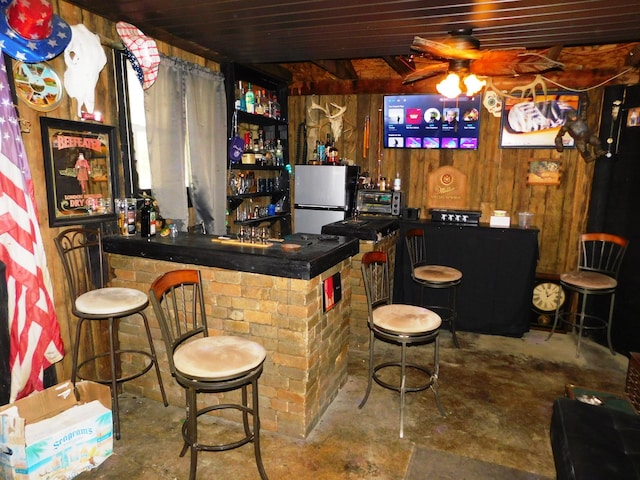 bar featuring freestanding refrigerator, wooden walls, a community bar, ceiling fan, and unfinished concrete floors