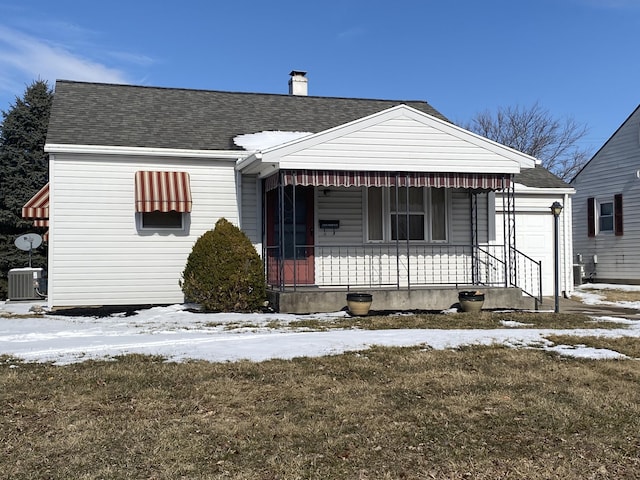 bungalow-style house with central AC unit, covered porch, a garage, a shingled roof, and a yard