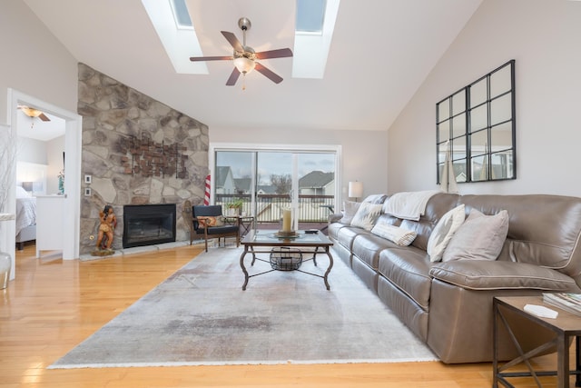 living room with ceiling fan, lofted ceiling with skylight, a stone fireplace, and light wood-type flooring